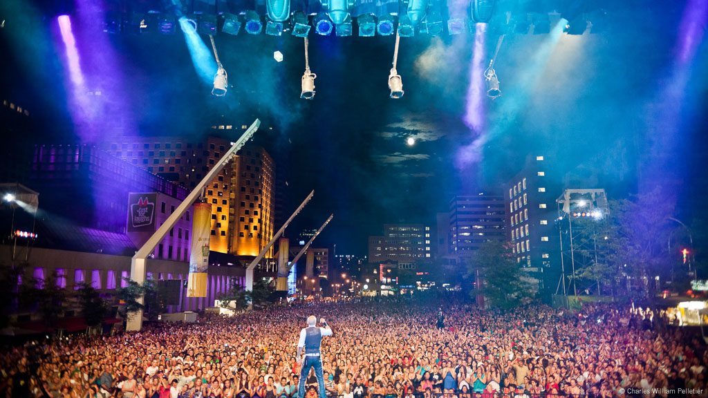 Une vaste foule de personnes se rassemble devant une scène illuminée de lumières bleues et violettes dans un cadre urbain nocturne.