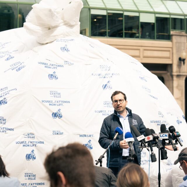 Glenn parlant derrière un pupitre, devant les journalistes