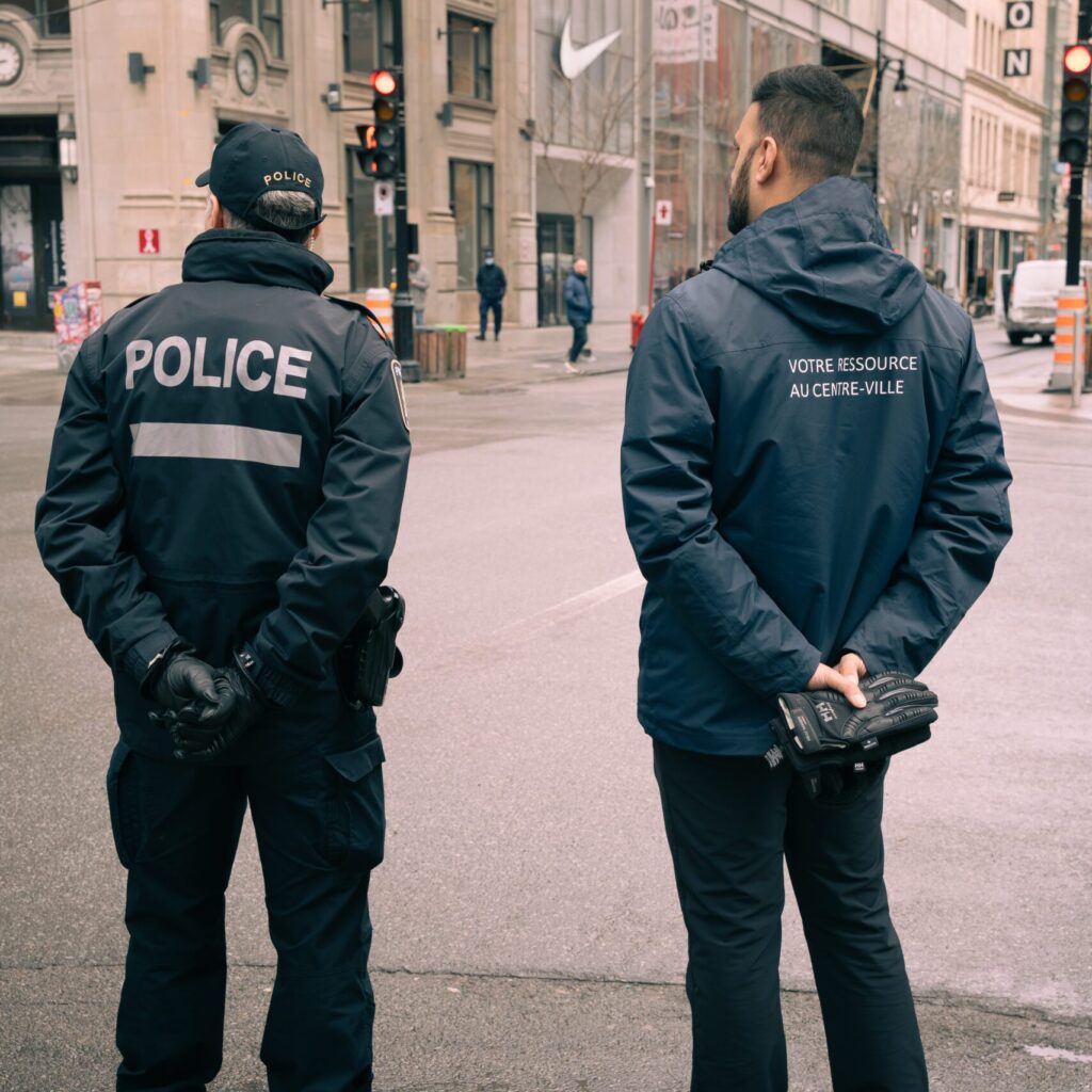 Un policier et un employé de Montréal centre-ville regardent la rue Sainte-Catherine, ils sont vus de dos.