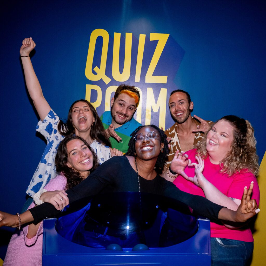 Un groupe d'amis souriant et enthousiaste sur un plateau de jeu Quiz Room, avec un mur bleu et le logo de l'entreprise.